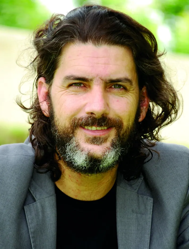 Retrato do celebrante Júlio César, de cabelo comprido e barba, sorrindo, com blazer cinza e fundo verde desfocado