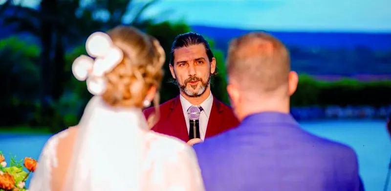 O celebrante fala ao microfone, de frente para a noiva e o noivo, numa cerimônia ao ar livre ao entardecer