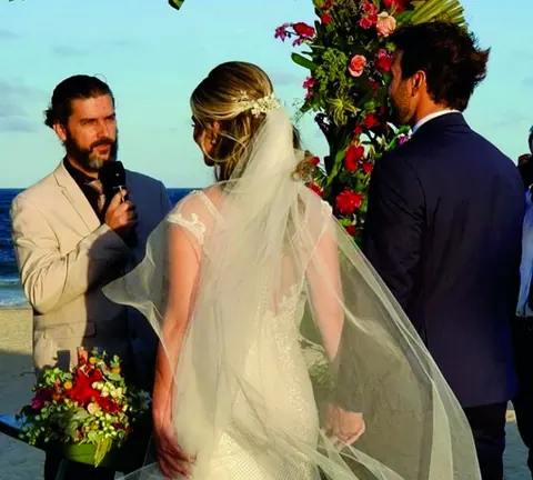 O celebrante fala ao microfone diante da noiva de véu e do noivo, junto a um arco de flores na praia